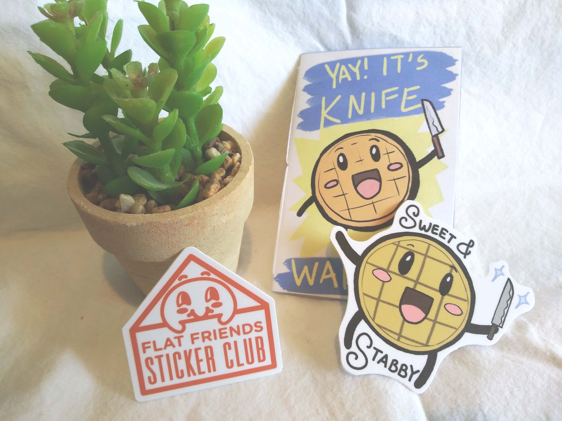 Two stickers and a mini booklet are next to a small potted succulent. The booklet says, "Yay! It's Knife Waffle!" and has a waffle with a cutesy face holding a knife. The sticker on the left is a logo of an enevelope with a cute, round face peeking out and the words, "Flat Friends Sticker Club" on it. The sticker on the left is the waffle with the knife and has the text "Sweet & Stabby" around the image