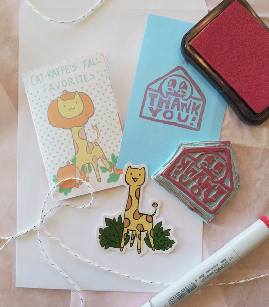 Image is a flat lay of several objects: a red marker, ink pad, a blue notecard that has been stamped with a stamp that says "thank you!", the stamp, a mini booklet, a sticker and some string. The booklet reads, "Catraffe's fall favorites" and shows a cat's head inside a pumpkin on a giraffe body. The sticker is the cat-raffe standing in some leaves and smiling. 