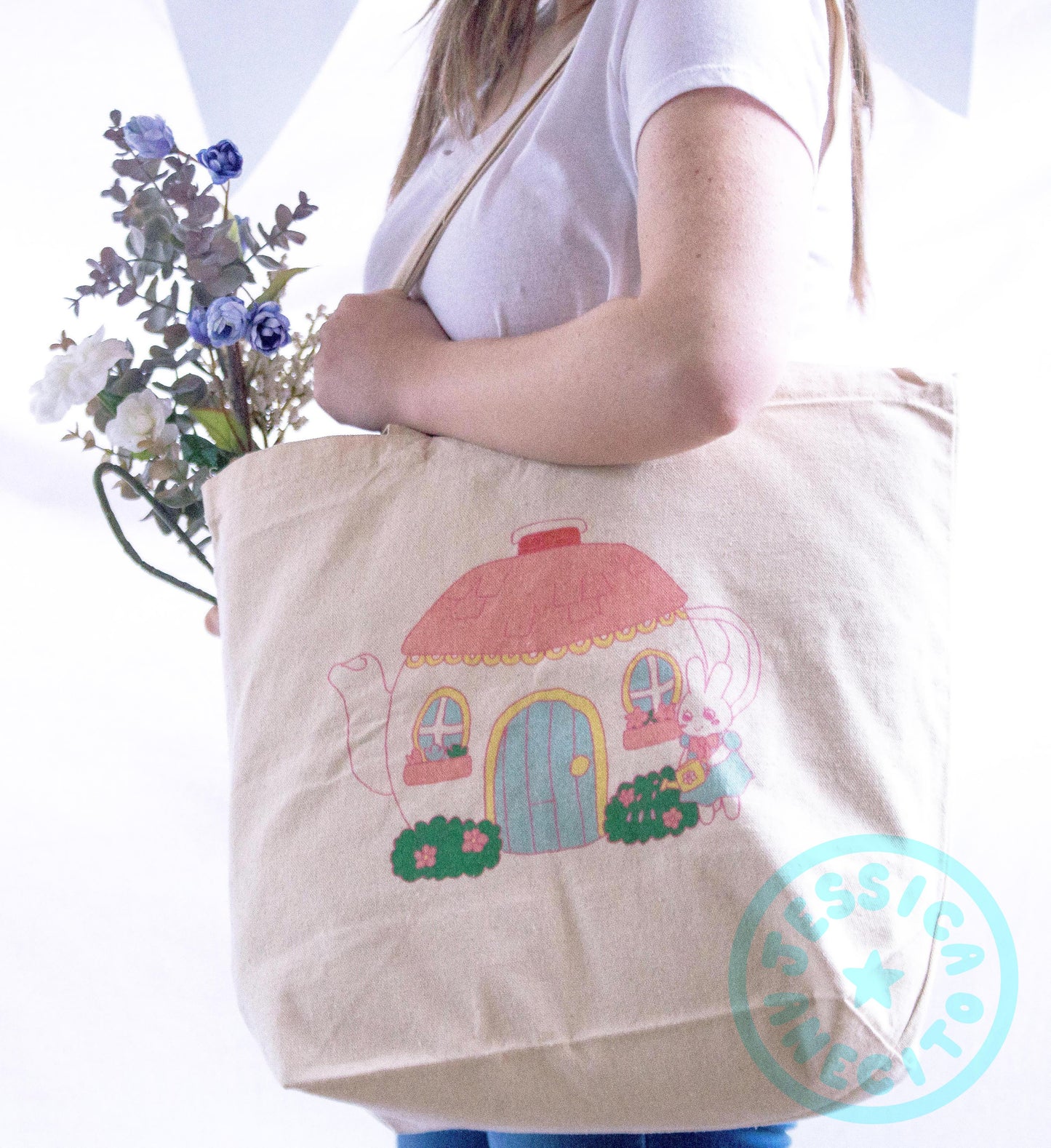 A female presenting person is standing at an angle holding a canvas tote bag over one shoulder. The bag is covering most of the body and has some purple and white flowers sticking out of it. On the side of the bag is an illustration of a bunny, standing like a person and wearing a blue dress with round sleeves, watering a bush. The bush has flowers on it and is front of a teapot that has been made into a house, with a door on the side and two small round windows. 