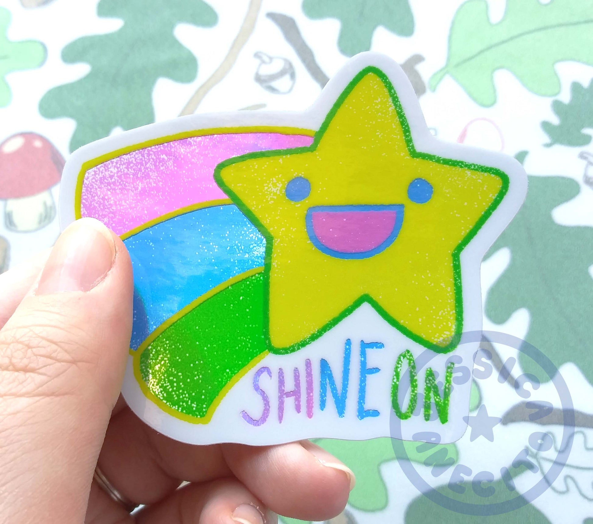 A hand is holding a shiny sticker of a shooting star. The star has a simple smiling face and three colors indicating it's a shooting star coming out of the left side in stripes. The stripes are pink, blue and green. Text under the star shape reads, "Shine on"