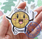 A hand is holding a sticker of a waffle with stick arms and legs smiling cutely and holding a knife. The knife has little sparkle drawings around it. The sticker reads, "sweet & stabby" 