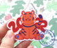 A hand is holding a sticker of a cute style tiger illustration. The tiger is smiling and sitting with his back legs spread on each side of his front legs, like a dog. The number 2022 is behind the tiger in red numbers. 