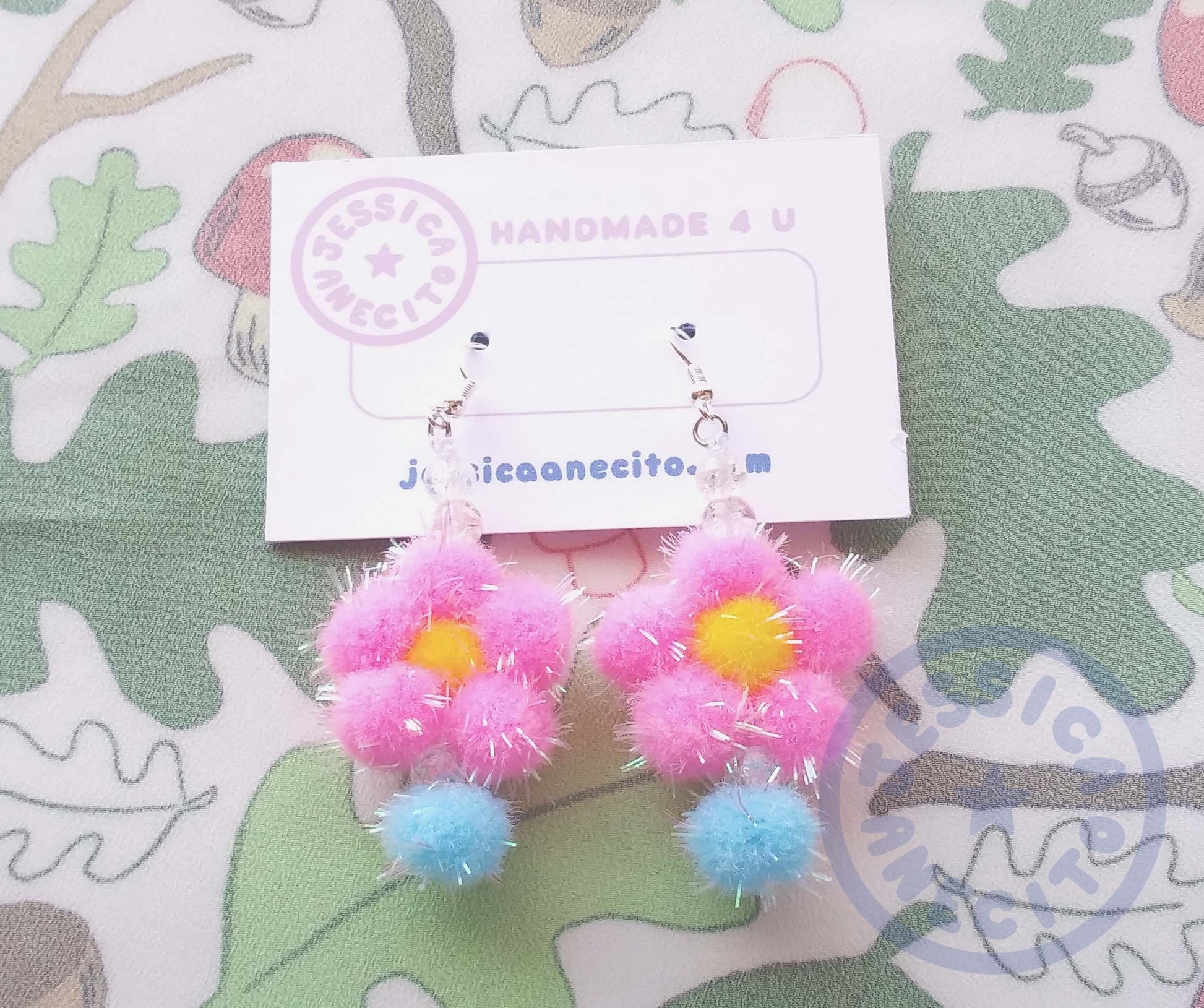 Two earrings are attached to a thick paper that reads, "Handmade 4 u" and "jessicaanecito.com". A logo is on the corner that is shaped like a circle and has the name "Jessica Anecito" going around the inside with a star in the center. The earrings are made of a yellow pom pom surrounded by five pink pom poms to make a flower shape. The flower has a clear bead and a blue pom pom hanging off the bottom and two clear beads on top connecting it to an earring hook. 