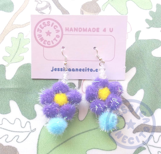 Two earrings are attached to a thick paper that reads, "Handmade 4 u" and "jessicaanecito.com". A logo is on the corner that is shaped like a circle and has the name "Jessica Anecito" going around the inside with a star in the center. The earrings are made of a yellow pom pom surrounded by five purple pom poms to make a flower shape. The flower has a clear bead and a blue pom pom hanging off the bottom and two clear beads on top connecting it to an earring hook.