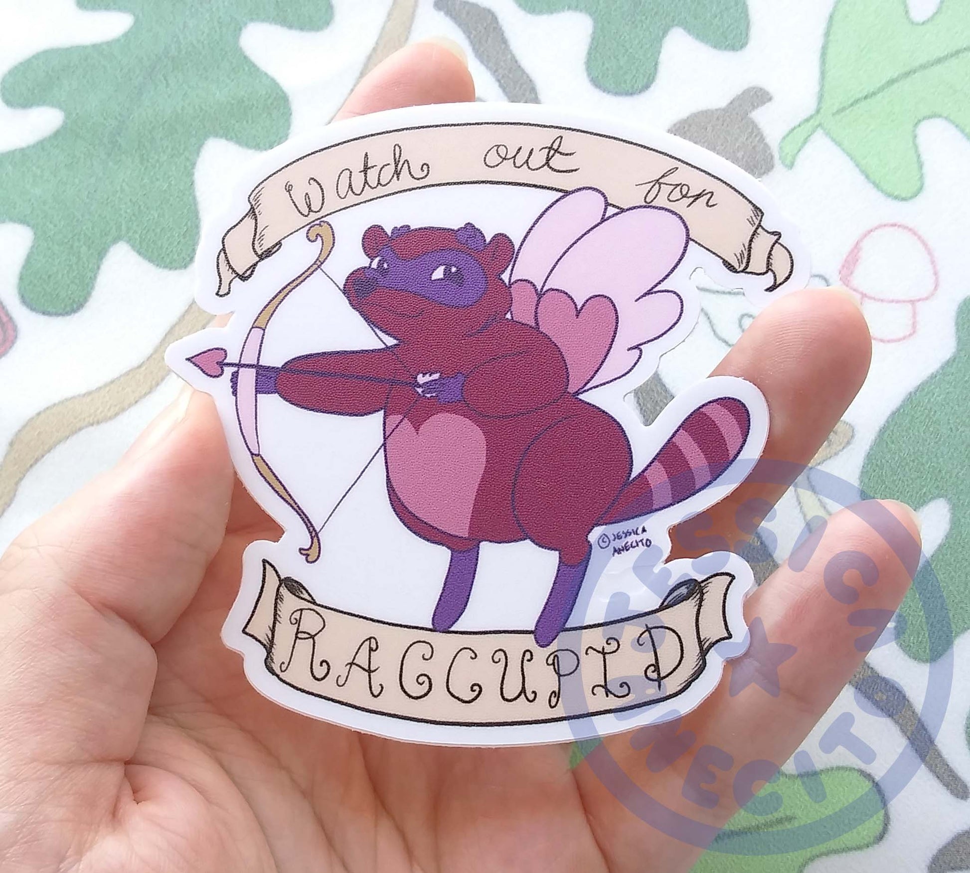 A hand is holding a smooth looking sticker of a purple and pink racoon. The racoon has a heart shape on it's belly and nose and pink wings. It is holding a bow with an arrow that has a heart instead of an arrowhead. Text on a scroll above and below the raccoon says, "watch out for Raccupid"